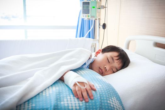 Illness asian boy sleeping at modern and comfortable equipped hospital room with saline intravenous (IV) on hand. Health care and people concept.
