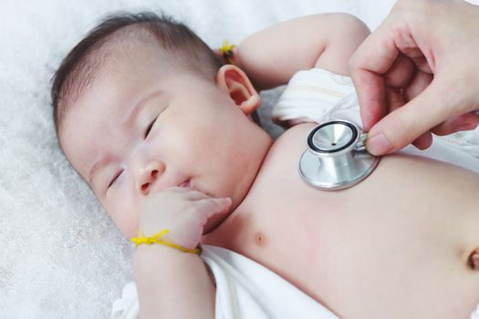 Professional pediatrician examining infant. Doctor using a stethoscope to listen to kid's chest checking heartbeat. Two months baby asian girl lying on sickbed in hospital.