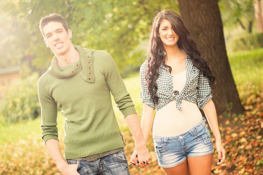 Young handsome heterosexual couple walking in park holding hands and smiling looking at the camera, while the sun shines from behind.