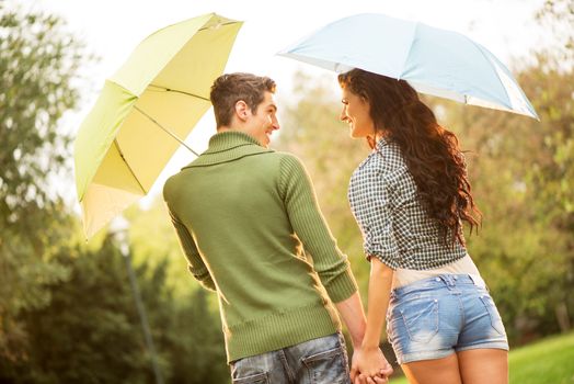 Rear view of a young couple in love with umbrellas while walking through the park holding hands and looking at each other with a smile.