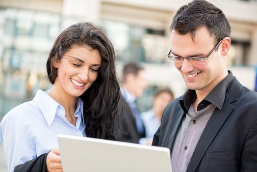 Smiling young businessman and businesswoman in front of office building looking at the laptop.