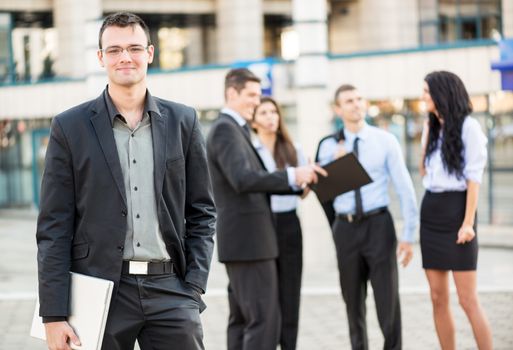 Young businessman with a laptop standing in front of office building separated from the rest of the business team. With a smile looking at the camera.