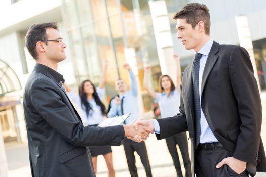 Two young businessman in front of office building shaking hands, while in the background there is a small group of business people hands raised celebrate the deal.