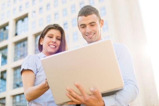 Smiling young businessman and businesswoman in front of office building looking at the laptop, while from behind the sun shines.