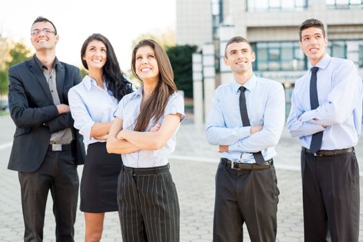 A small group of young business people standing in front of office building with arms crossed and with a smile on their faces looking in height.