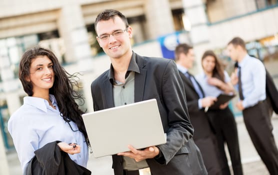 Young businessman with a laptop and businesswoman standing in front of office building separated from the rest of the business team. With a smile looking at the camera.