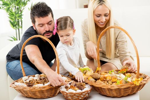 Happy family together, parents with daughter sitting on the couch in the living room in front of woven baskets filled with pastry, choosing the next snack.