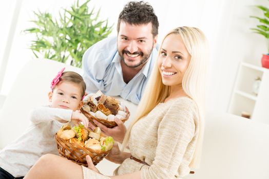 Young parents hold in their hands woven basket with delicious salty and sweet pastries. Beside them sit on the couch their little daughter eating pastries.