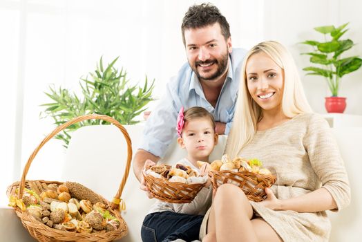 Young parents hold in their hands woven basket with delicious salty and sweet pastries. Beside them sit on the couch their little daughter and eating pastries.