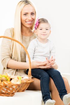 Beautiful blonde young mother with her cute little daughter, who is sitting in her lap, smiling looking at camera. Beside them on the couch are woven baskets with pastries.