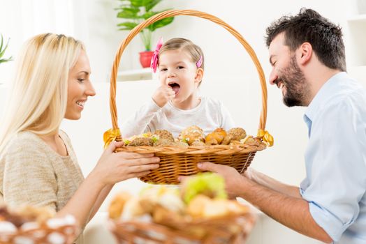 Young parents kneel in front of their little daughter with a smile watching pastries from a large woven basket which together hold in their hands.