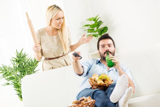 Young man sitting on a couch, holding a bottle of beer in one hand, remote control in the other hand,on the knee had baskets with pastries, while behind him stands a young woman with a rolling pin in hand with an angry expression.