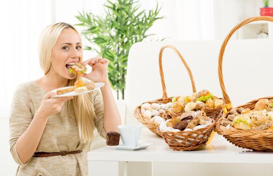Young pretty blonde woman eating a small sandwich, in the background you can see woven basket with bakery products. With an expression of enjoyment on her face looking at the camera.