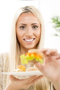 Beautiful young blonde woman with a smile on her face looking at the camera, holding a mini sandwich that is out of focus, offering the viewer a bit.