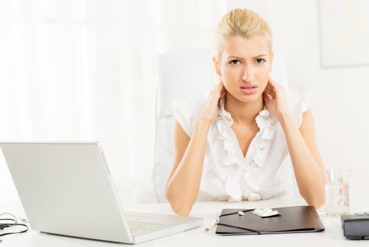 Young businesswoman at work, holding her neck with a pained expression on her face looking at the camera. In front of her on her office desk are laptop and pills.