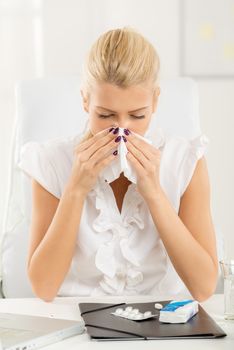 A young blonde business woman at her workplace at an office desk in front of laptop, shaking her nose into a handkerchief ready to take the pills.