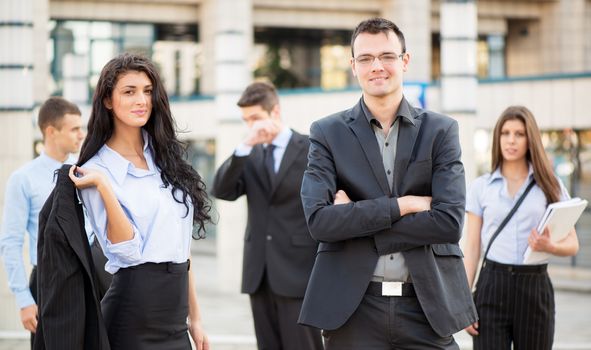 Young businesswoman and businessman, with their team in the background, standing in front of office building, with a smile looking at camera.