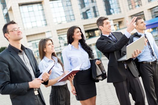 Young businessman with laptop in front of office building outstretched fingers of the hand, aimed at heights, explaining his colleagues business plan.