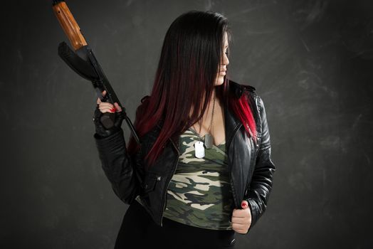 Profile of an attractive young woman in a leather jacket and camouflage shirt with cleavage with a Kalashnikov in her hand.