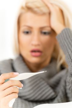 Young pretty blonde girl, out of focus, worriedly looking at a digital thermometer that is in the foreground.