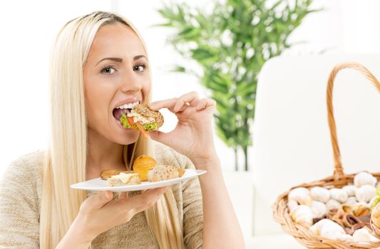 Young pretty blonde woman eating a small sandwich,with an expression of enjoyment on her face looking at the camera.
