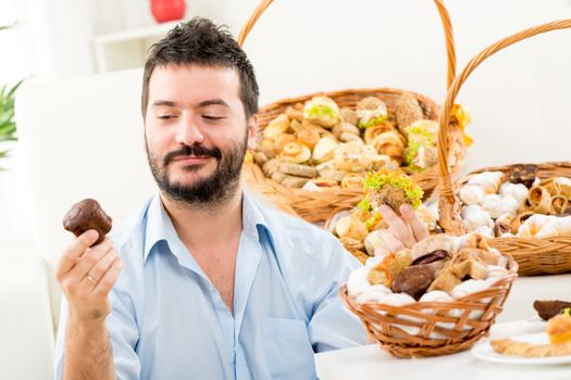 Young man with a beard, holding in his hands a sweet and savory pastries. With a smile looking at sweet cake. In the background you can see woven baskets with pastries.