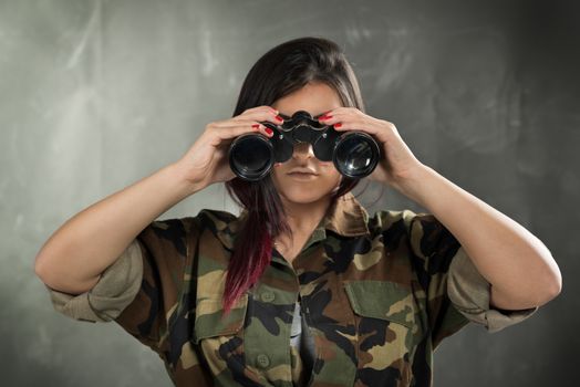 Young woman wearing a camouflage uniform, looking at the camera through binoculars.