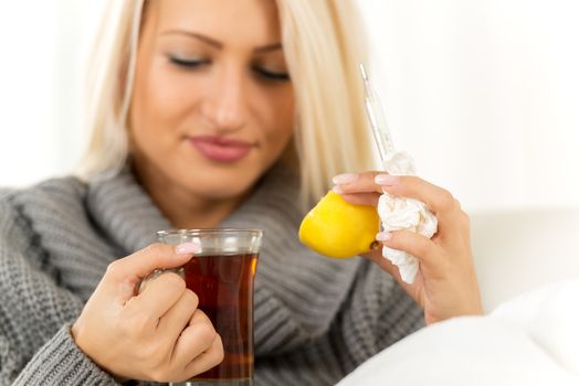 Blonde girl with face out of focus holding in one hand a cup of tea, and in the other lemon, thermometer and a crumpled handkerchief. In the foreground are tea and lemon.
