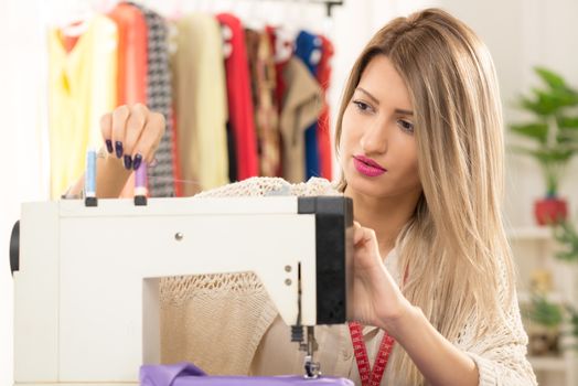 Young beautiful woman sitting in front of a sewing machine on which sets the thread, in the background you can see hanging garments.