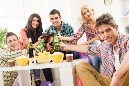 Cheerful friends at home party toasting knocking with bottles and glasses. With a smile looking at the camera.