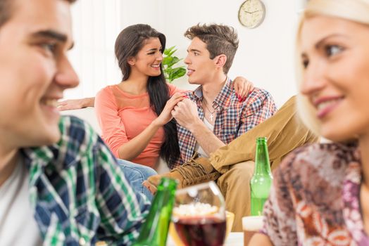 Young couple at a house party, sit on the floor, behind them is another young couple which is in the foreground, sitting on the couch and look at each other.