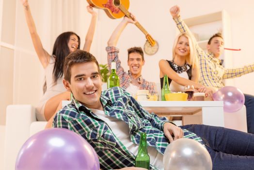 Young guy at house party, lying on the floor, smiling, looking at the camera in the background you can see his friends sitting on the couch happy with their hands raised.