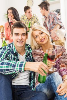 Young couple at a house party, sit on the floor and knocking with drinks, with a smile looking at the camera. In the background you can see their friends who are sitting on the couch.