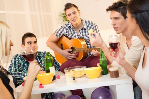 Small group of young people at home party,  enjoying the time together with music, drinks and snacks. In the foreground is a guy with an acoustic guitar, with a smile looking at the camera.