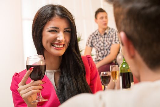 Young pretty brunette girl with a glass of wine in hand, with a smile looking at the guy who has his back turned to the camera, while in the background see another young guy in the ambient house party.