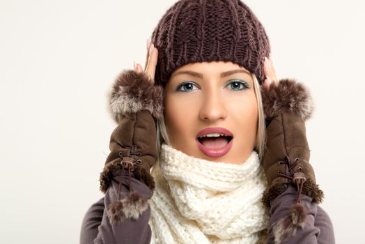 Beautiful young blue-eyed woman with woolen hat and fur gloves, wrapped in a scarf, holding her head with an expression of surprise with her mouth open looking at the camera.