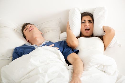 Heterosexual couple in bed, man sleeps and snoring with mouth open, while a woman irritated by snoring covers the ears with a pillow.