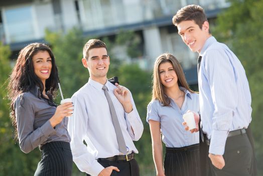 Group of young business people standing outside near their company on the coffee break, enjoying the beautiful day and with a smile on their faces looking at the camera.