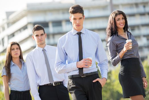 Young businessman, elegantly dressed, standing  with his team on a coffee break in front of office building and looking at the camera with a serious expression on his face.