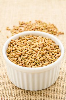 Fenugreek seeds in white bowl on sack background.