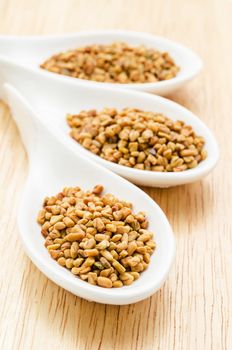 Fenugreek seeds in white spoon on wood background.