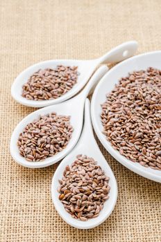 Brown linseed or flax seeds in white spoon on sack background.