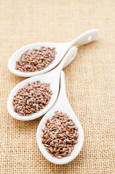 Brown flax seeds or linseed in white spoons on sack background.