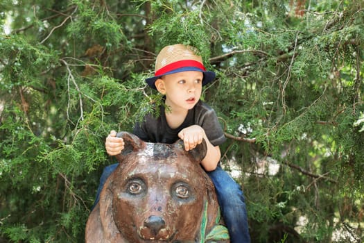 Cute little boy wearing hat playing on the playground in the summer in the city. Happy boy sitting on the bear statue in zoo