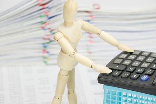 Wooden dummy using calculator with blue basket on finance account have blur pile overload document of report with colorful paperclip as background.