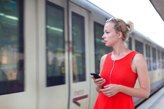 Young woman in red dress with a cell phone in her hand waiting on the platform of a railway station for their train. to arrive.