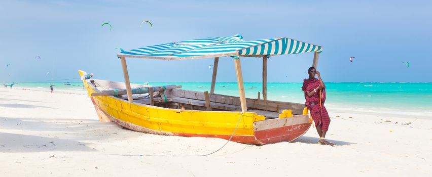 Maasai warrior lounging aroundon traditional colorful wooden boat on picture perfect tropical sandy beach on Zanzibar, Tanzania, East Africa. Kiteboarding spot on Paje beach.