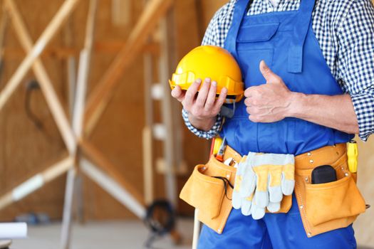 Builder holding hardhat with thumb up at construstion site