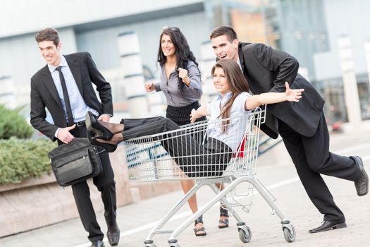 Group of cheerful young business people, elegantly dressed, in motion rush, push trolley for shopping, in which sits a young businesswoman.