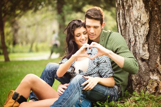 Young heterosexual couple sitting on the grass in the park next to the tree and touching with the fingers forming a heart shape.